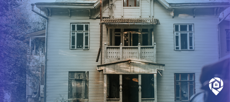 Weathered two-story house with boarded windows and a damaged porch, the kind of distressed property investors scout for deals.