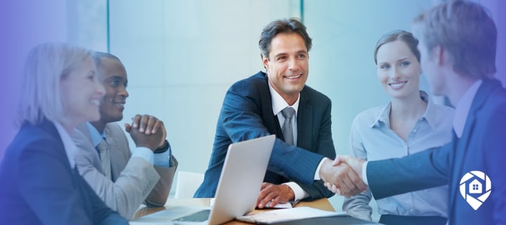 A team of business professionals at a conference table, two shaking hands over a laptop.